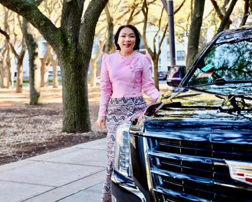 Woman in a pink traditional-style top standing beside a shiny black SUV in a tree-lined park.